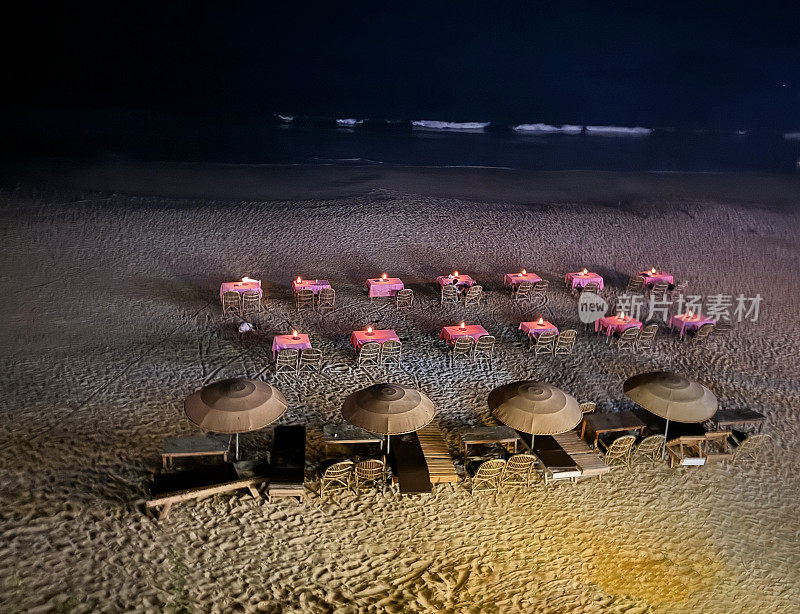 海滩餐厅夜景特写，印度果阿，帕洛莱姆海滩和阿拉伯海，一排排铺着桌布和椅子的竹桌，为露天用餐做好了准备，点燃的蜡烛桌中心，日光浴椅，遮阳伞