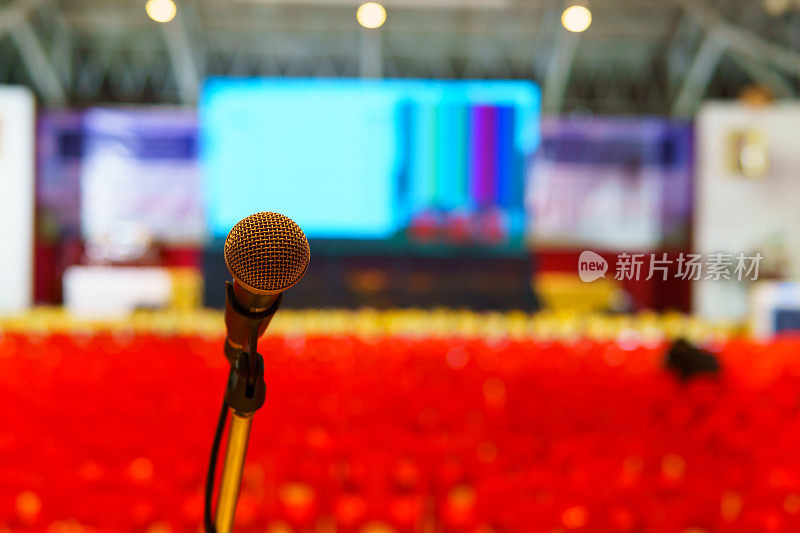 近距离的麦克风在研讨会大厅扬声器或歌手模糊排红色塑料椅子和大led显示屏背景。为文本复制空间。讲师、演讲者、研讨会概念。