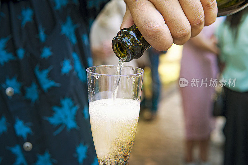 这是一个男子在户外节日活动中举起的半满香槟酒杯的特写。在模糊的背景下，男嘉宾往玻璃杯里倒满了起泡酒。婚礼的传统。