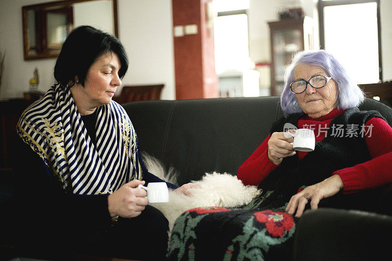 漂亮的老妈妈和她的成年女儿拿着杯子，坐在家里的沙发上聊天