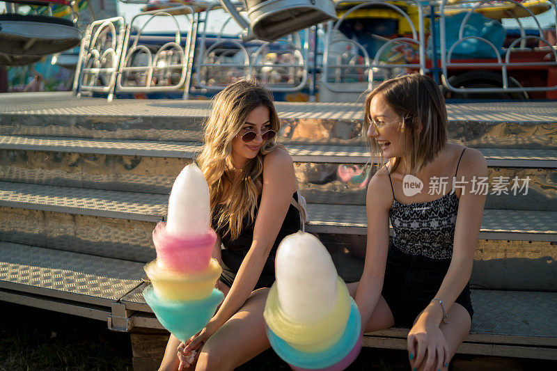 快乐的女性朋友在游乐园吃棉花糖的照片。两个年轻女人在游乐园玩了一天。