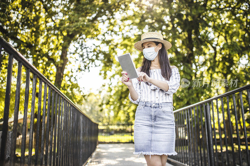 时尚年轻女性在冠状病毒大流行期间戴着医用防护口罩放松、使用平板电脑和在自然中度过时光的肖像