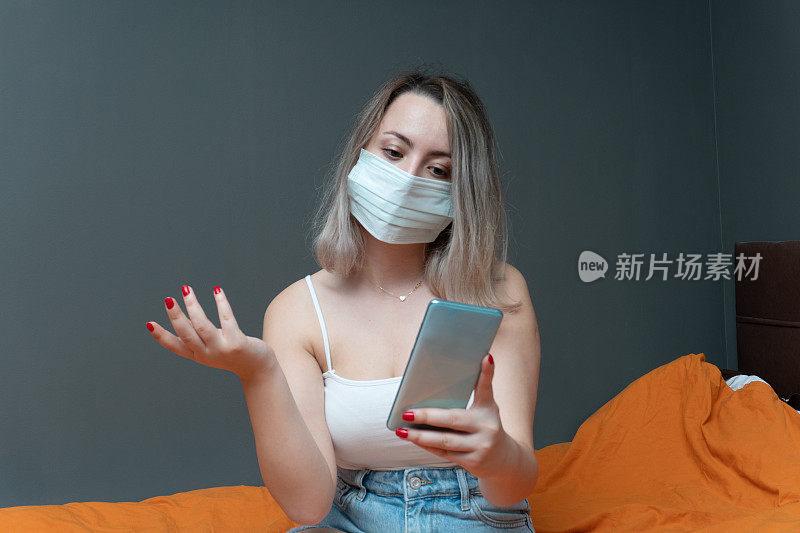 年轻美丽的女人用智能手机戴着外科口罩在床上的肖像