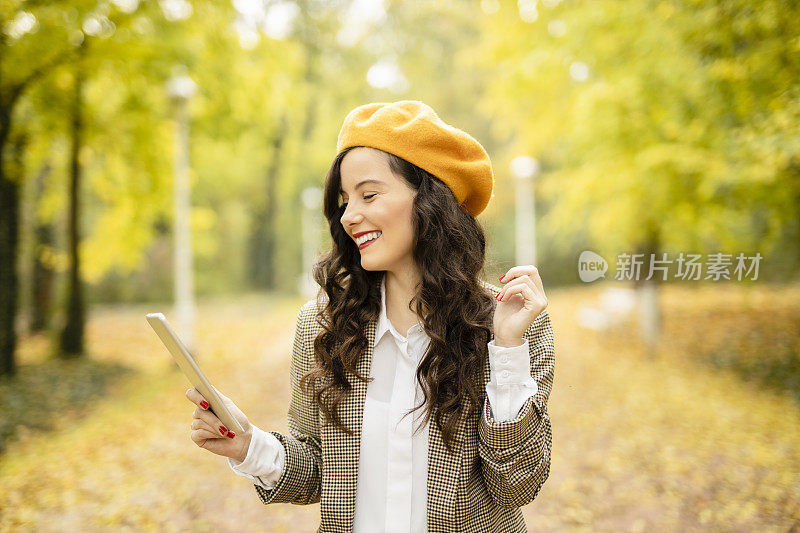 美丽的年轻女子在她的数字平板电脑上阅读杂志或报纸，享受秋天的公园