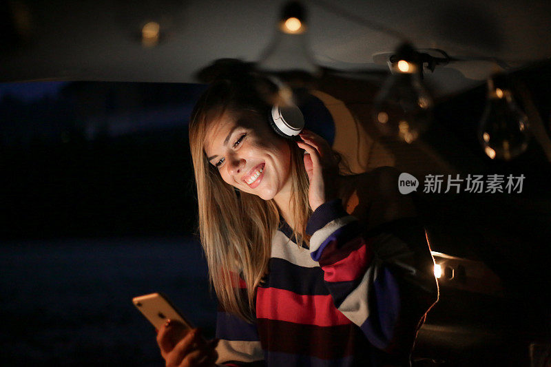 一个年轻漂亮的女人露营在车的后面，一边放松一边用她的手机和耳机听音乐