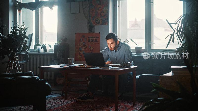 年轻英俊的男子坐在沙发上，工作笔记本电脑在创意机构在真实的Loft办公室雨天。装修时尚的装饰与室内植物，艺术海报和窗户。