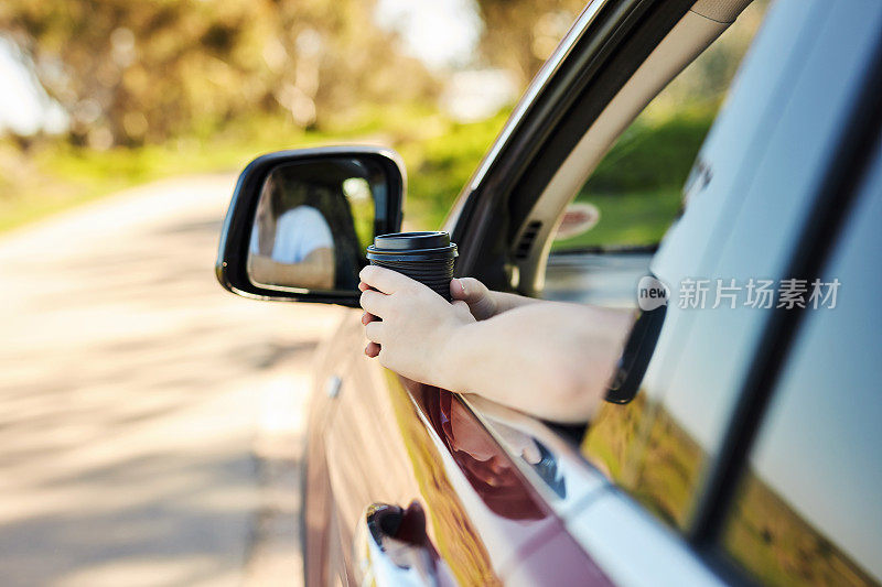 一个女人在路上开车，她的左臂伸出车窗，拿着一个咖啡杯。手拿着咖啡纸杯站在车窗外。