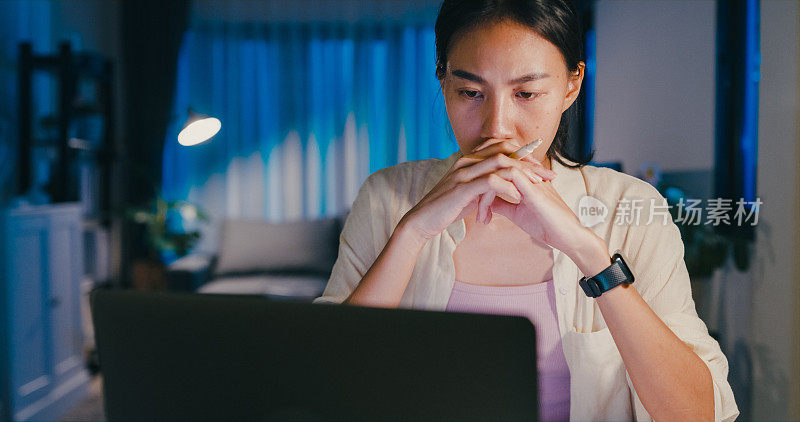 年轻的亚洲女性穿着正装衬衫坐在桌子前，拿着笔记本电脑思考和得到的想法，使紧急报告会议加班到晚上在家。