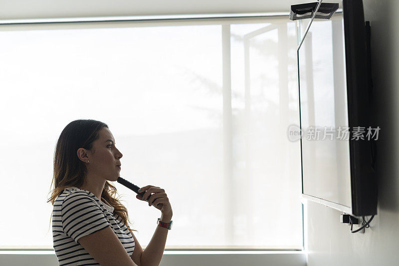 一位拉丁妇女站在电视机前看电视剧，手里拿着遥控器