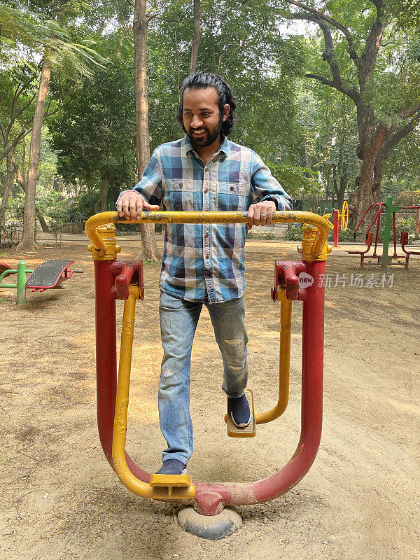 穿着牛仔裤和衬衫的印度男子在户外健身房使用空气行者健身运动设备，在户外露天公园锻炼