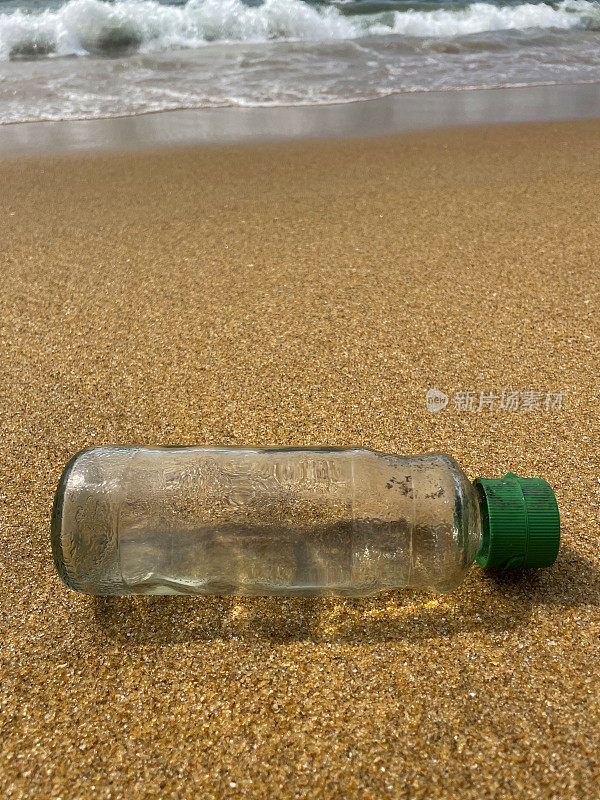 特写图像，空的，透明的塑料饮料瓶与绿色盖子被冲到沙滩上的退潮水的边缘，破碎的海浪冲刷海洋垃圾和岸上的污染，乱扔的沙子，肮脏的海滩，关注前景，复制空间