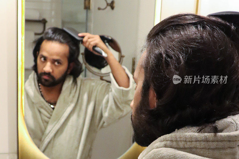 镜子里的全画幅印度男人穿着浴袍站在浴室水槽前，用梳子梳理凌乱潮湿的头发，自我护理和修饰的概念