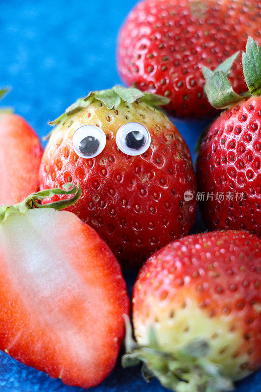 有趣的全画幅图像，由成熟的整颗草莓制成的卡通人物，新鲜的草莓和大眼睛围绕着整颗和半颗的草莓果实，蓝色的背景，抬高的视角，儿童健康饮食，幽默的概念
