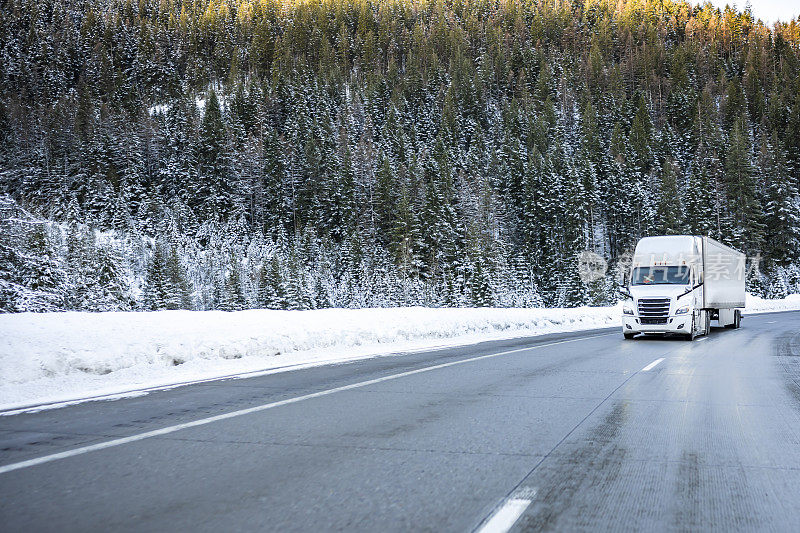 长途白色大钻机半挂车，用干式货车半挂车运输货物，在冬季湿滑的结冰公路上爬坡，路肩上有雪，山上有森林