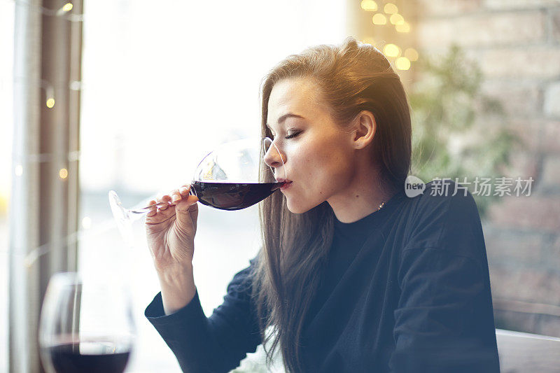 美丽的年轻女子喝红酒与朋友在咖啡馆，肖像与葡萄酒杯靠近窗户。假期假期晚上概念