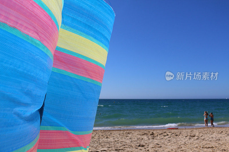 色彩鲜艳的条纹防风林在沙滩上随风翻滚，背景中海浪拍打着水的边缘，聚焦于前景