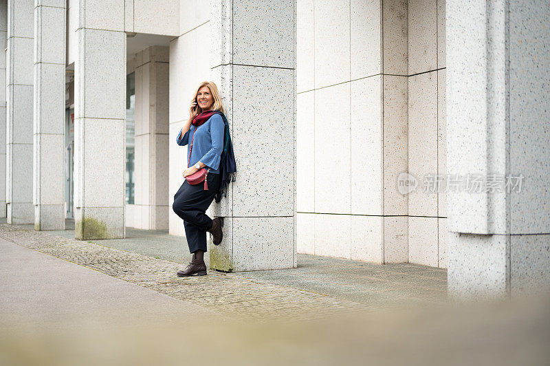 成年快活的女人靠在一根建筑柱子上，在户外用手机聊天