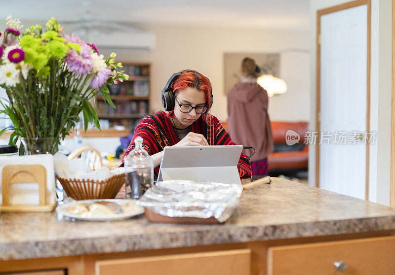 在家学习并不总是舒适的。这个十几岁的女孩戴着耳机在远处上课，在厨房柜台边用笔记本电脑工作，这时她的大姐正从后面走进客厅。