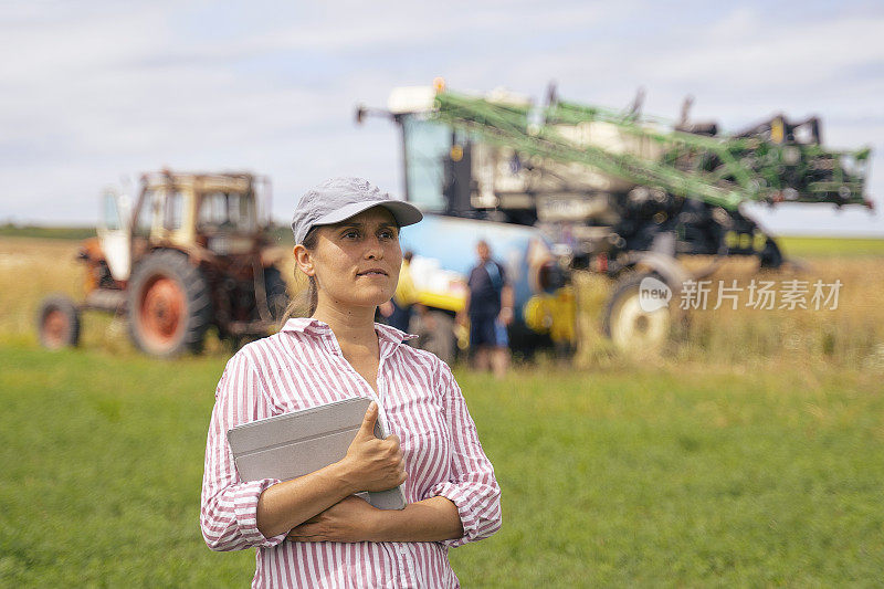 一个年轻的农妇在田地里工作的肖像，在喷洒庄稼的时候操作一个现代的作物喷雾器。在一个阳光明媚的日子里，有机农场的收获季节。农作物喷雾器使用远程控制。