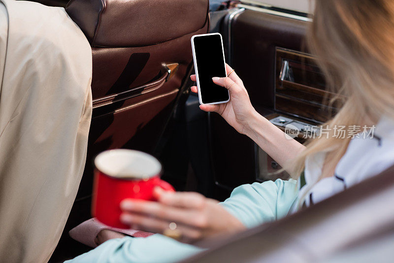 女性抱着一杯咖啡和智能手机的空白屏幕，而坐在cabriolet模糊的前景