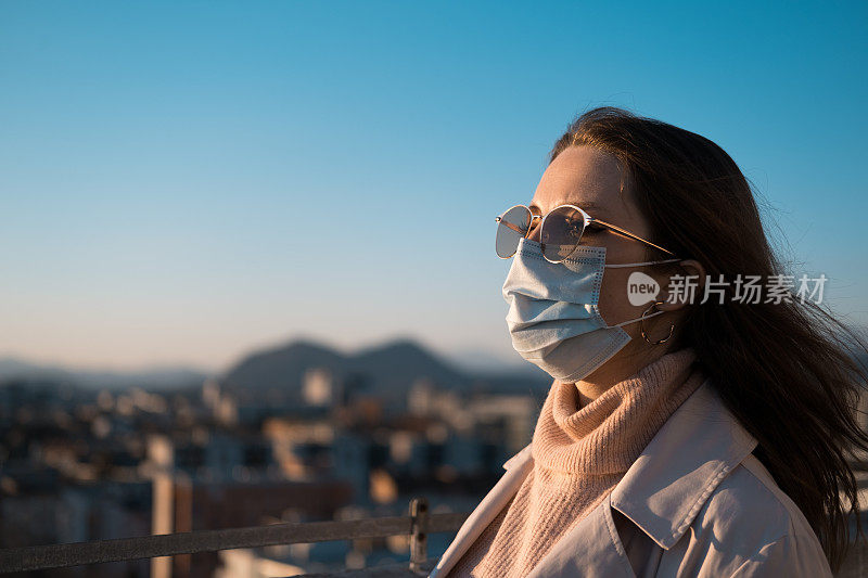 风吹着头发的女人戴着医用口罩和太阳镜，站在外面，看这个城市。