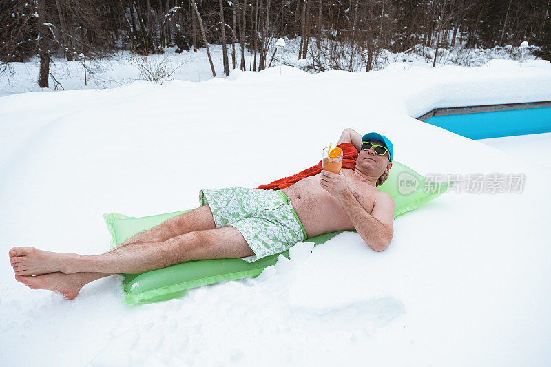 一个穿着泳裤的年轻人躺在充气床垫上，手里拿着一杯鸡尾酒。一个男人正在一个覆盖着雪的室外游泳池旁休息。