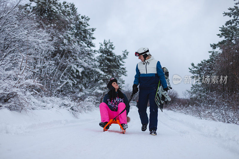 年轻的男子滑雪板拉着他的女朋友在雪橇上，他的滑雪板在下雪的环境