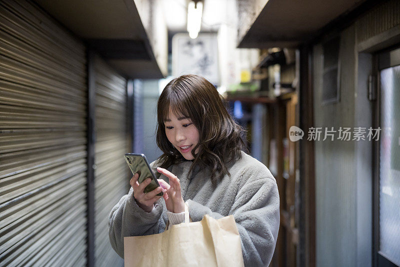 女人站在狭窄的购物街上，用着智能手机