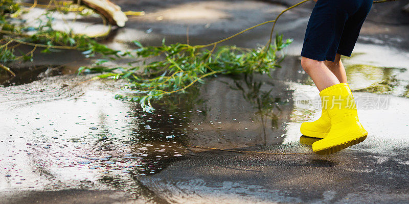 夏雨。水坑在沥青。穿着黄色雨靴的孩子。孩子跑过水坑。寒冷的秋天的雨。潮湿的天气。水飞溅。倒影在水中
