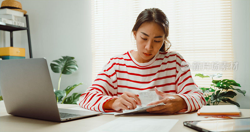 年轻的亚洲女人穿着毛衣坐在书桌前用笔记本电脑使用计算机计算器水电费账单计算检查信用卡收据每月的费用账单在家里。