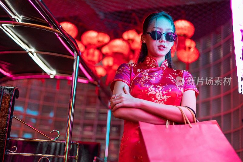 一位年轻的亚洲女模特肖像照片，戴着太阳镜，穿着红色中国传统旗袍，在霓虹灯前看起来很酷，拿着购物袋在一辆嘟嘟出租车旁边