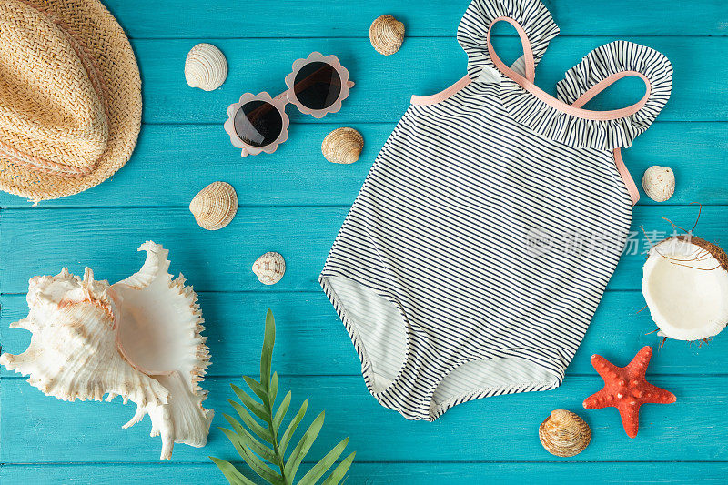 儿童连体泳衣和草帽旁边的海滩配件，贝壳，海星和椰子上的木制蓝绿色背景。夏日氛围和海边度假的概念。平躺