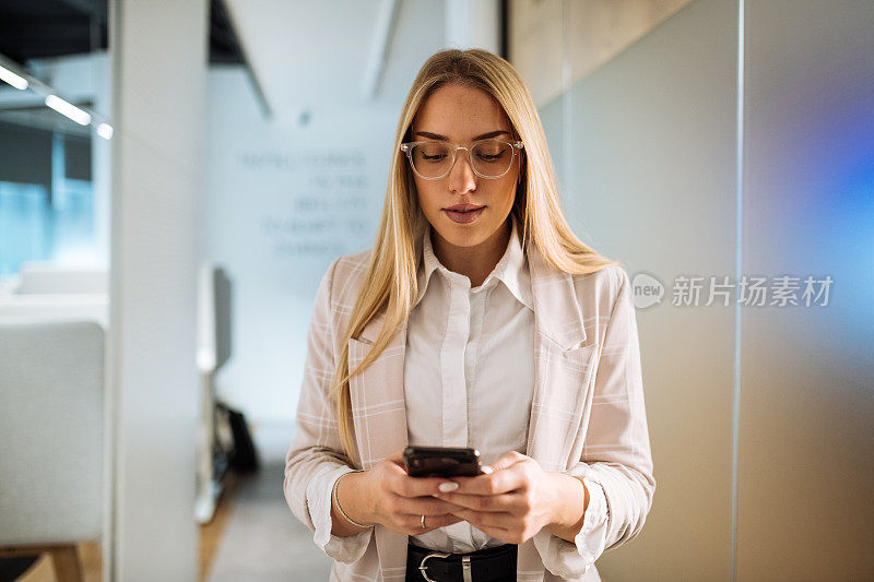 坚定的年轻女商人，穿着正装，在办公室工作，在大厅走动，使用智能手机