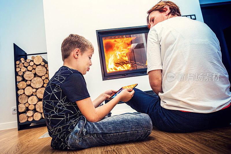 母亲和儿子坐在家里的壁炉前，儿子正在使用平板电脑