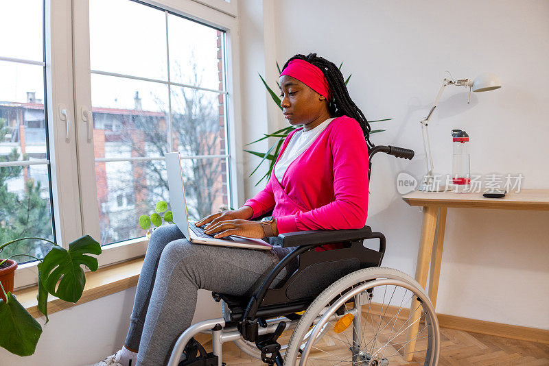 黑人妇女坐在轮椅上用笔记本电脑工作，靠窗