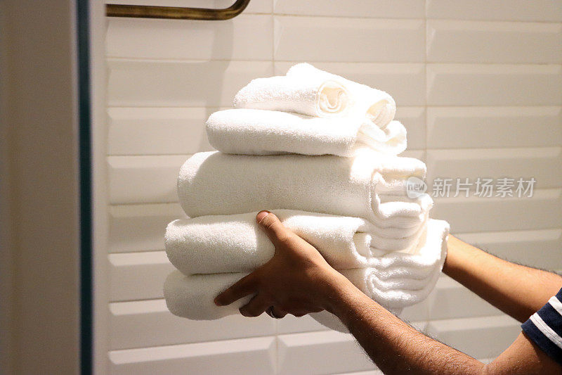 特写图像，一个无法辨认的人拿着一堆白色的酒店毛巾，折叠的浴巾和手巾被放在高架子上，豪华的酒店浴室有白色斜边墙砖在水平堆叠模式，关注前景