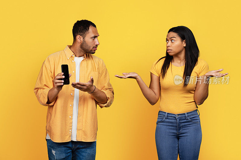 悲伤的严肃的年轻非洲裔美国人展示电话给困惑的女人孤立在黄色背景