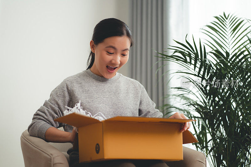 亚洲女人打开纸箱，开心兴奋地往里看，坐在家里客厅的扶手椅上。