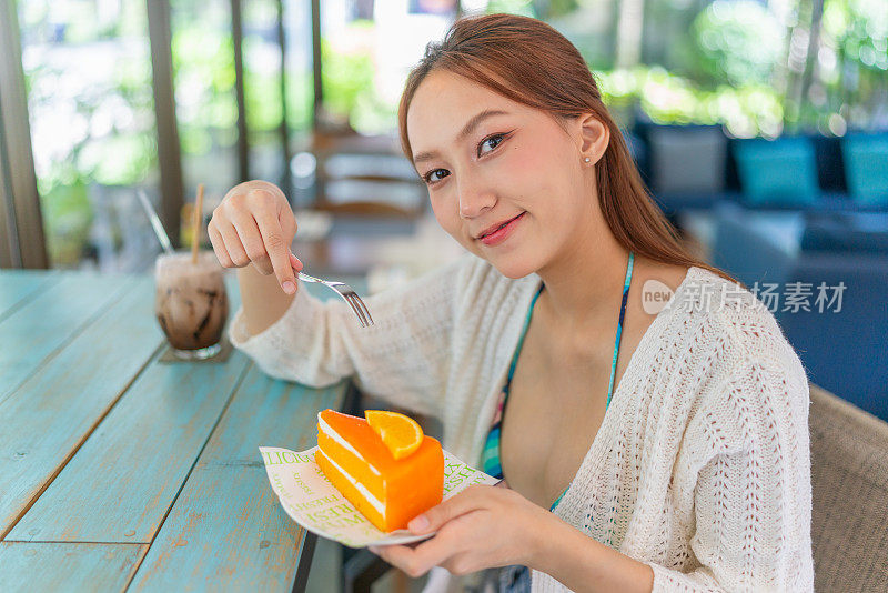 年轻的亚洲女子在海滩咖啡馆喝着冰拿铁，一边享用着一片橘子蛋糕。穿着沙滩装的女性数字游牧民在度假氛围中休息。穿着沙滩装的亚洲女人在咖啡馆里放松。