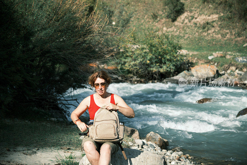一位女游客坐在山河边，翻看她的背包