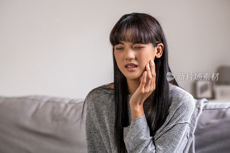 一名年轻的亚洲女性，明显感到疼痛，捂着脸颊，这表明她可能牙痛或牙齿敏感。