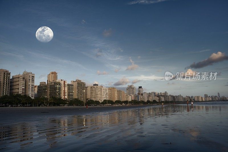 巴西桑托斯市。下午晚些时候，桑托斯海滩上的海边建筑被太阳照亮，蓝天上挂着一轮满月。
