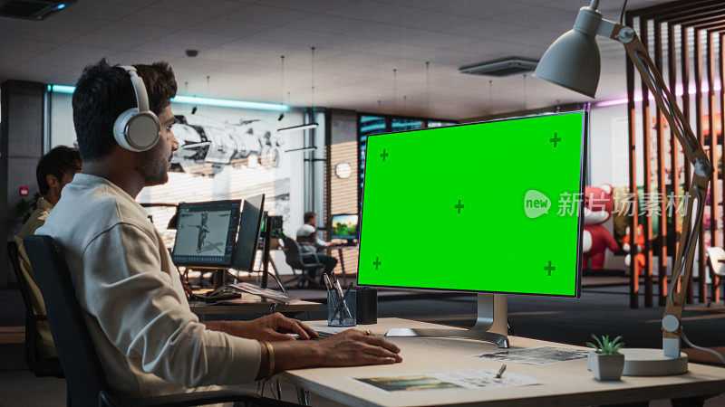 印度男性游戏设计师使用带有绿屏Chromakey的桌面电脑，在3D建模软件中为新视频游戏设计沉浸式世界。在游戏开发办公室工作的人