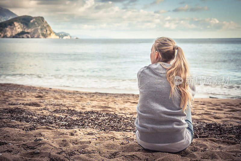 一个忧郁孤独的年轻女子坐在海滩上，抱着膝盖，满怀希望地望着远方