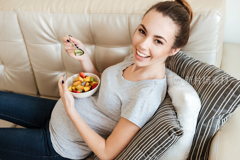 快乐的孕妇坐在沙发上吃水果沙拉