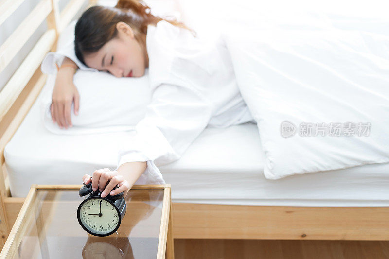年轻的亚洲女子按下贪睡的按钮，早上醒来闹钟的警报和睡在卧室旁边的懒人在家