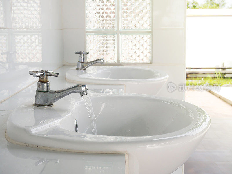 水从水龙头流出。公共浴室清洁卫生的概念。带有白色水槽的金属龙头。