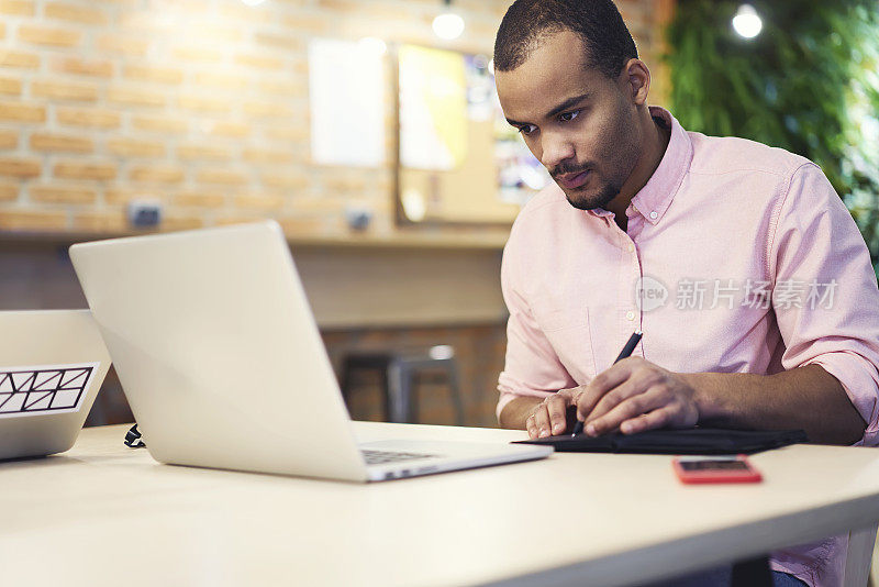 熟练的专注的非裔美国平面设计师坐在办公室的笔记本电脑上，一边观看办公大楼的在线方案，一边在笔记本上绘制创意规划的草图