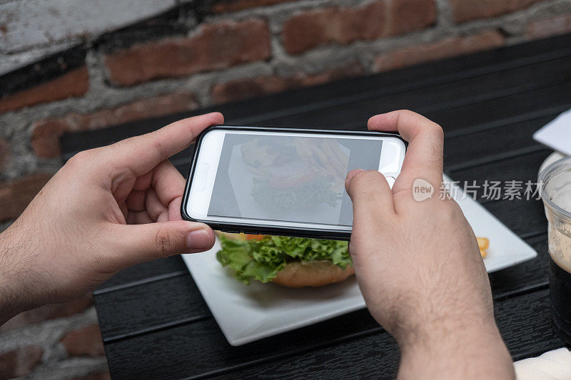 一名男子拿着手机给他的食物拍照。智能手机食品摄影。在一家户外酒吧给汉堡和炸薯条拍照。