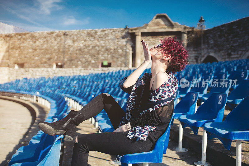 迷人的红发女人坐在空旷的露天剧场，享受在阳光明媚的日子。